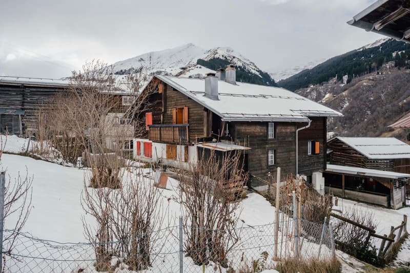 Mountain house with wooden facade, covered in snow, chimneys, two windows with red shutters, and mountain backdrop