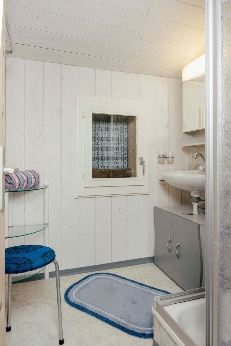 White walls, white sink, gray cabinets, window, rug, chair, mirror