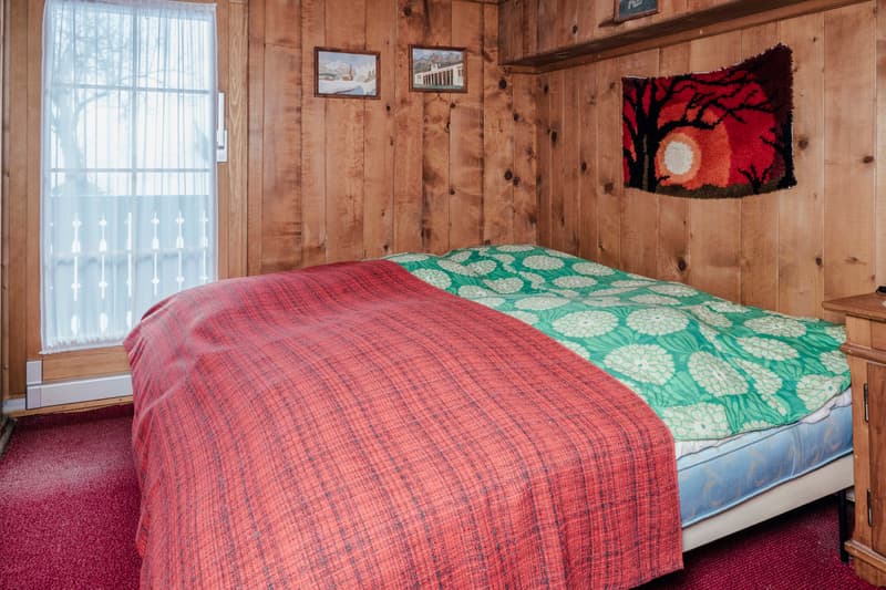 bedroom with wooden walls, white curtains, wooden nightstand, two pictures on wall, one rug on wall, bed with a red blanket and green bedspread
