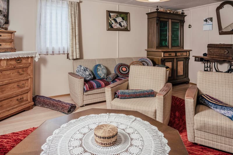 Wooden dresser, sofa, two armchairs, carpet, coffee table with doily, rug, cabinet, picture frames, mirror, calendar, rug, woven basket, throw pillows, throw blanket