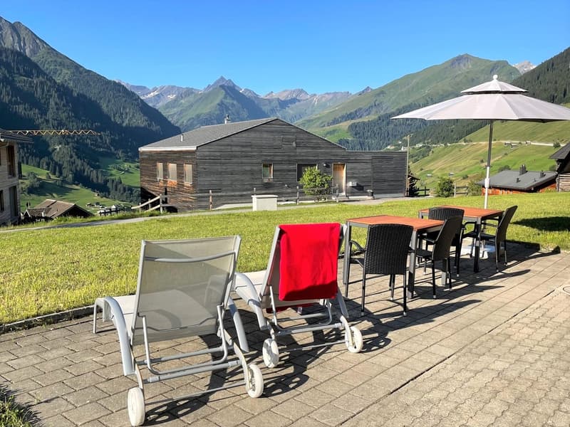 Outdoor seating area with chairs, table, and umbrella, surrounded by a grassy field, with a wooden house and mountains in the background.