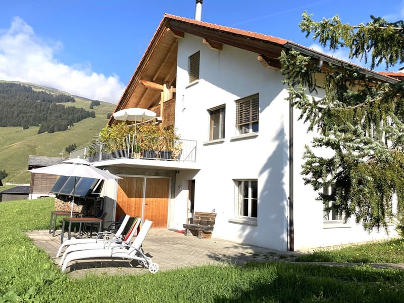 2 story house, white exterior, brown wooden door, balcony with railings, outdoor seating area with umbrellas and lounge chairs, surrounded by grass and trees, mountain view