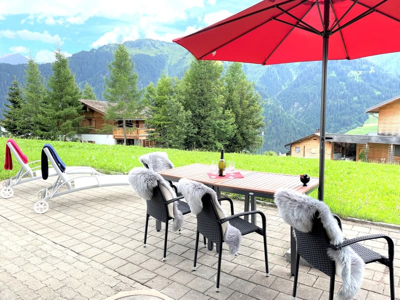 Outdoor dining table with chairs, red umbrella, green grass, mountains in the background