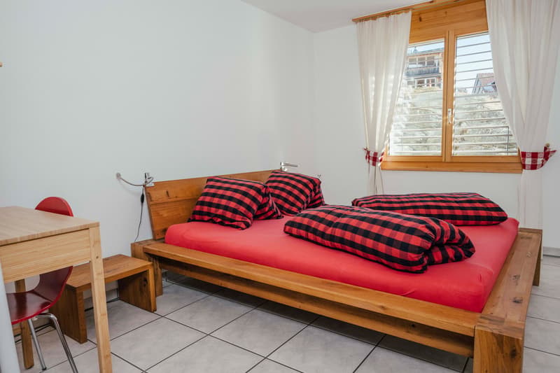 Bedroom with wooden bed, red bedspread, black and red checkered pillows, wooden desk, red chair, white walls, tiled floor, window with white curtains