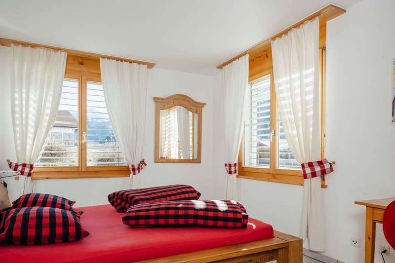 bedroom with wooden bed, red bedsheet, red and black checkered pillows, wooden mirror, wooden windows, white curtains