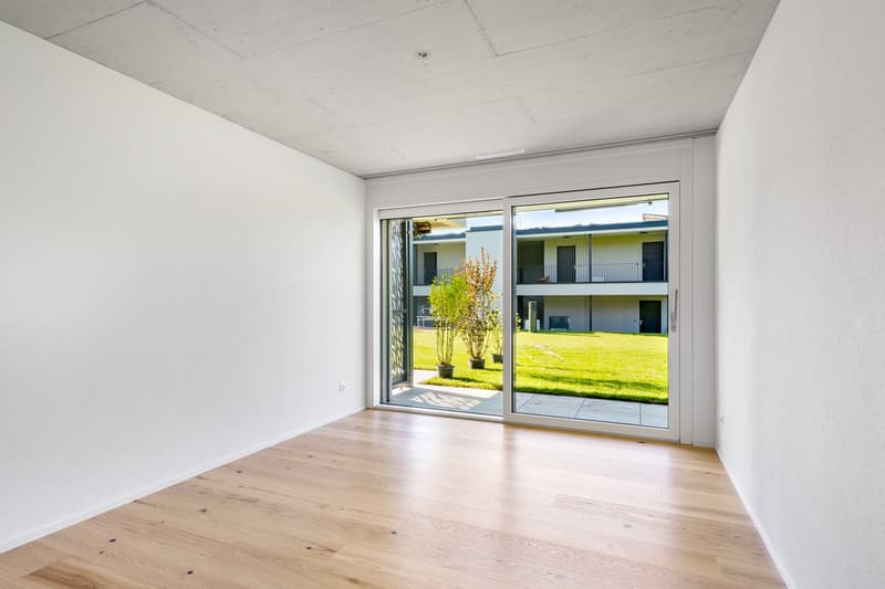 Sliding glass door leading to a balcony, hardwood floor, white walls, empty room, glass doors with a view of a grassy area and another building.
