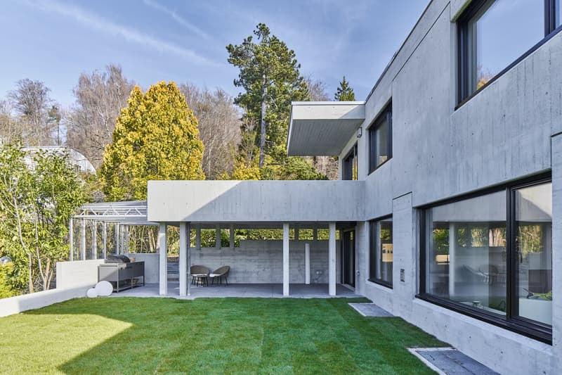 Modern concrete house, large windows, outdoor seating area, grill, well-manicured lawn, trees