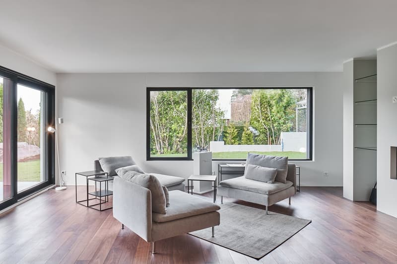modern living room with large windows, wooden floor, gray couches, side tables, rug, and a view of the garden