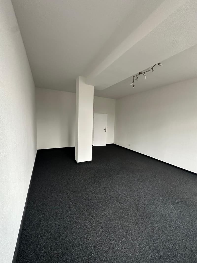 Empty room with white walls, black carpeted floor, and track lighting on the ceiling