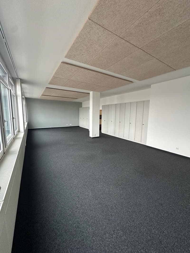 The image shows a large open office space with a gray carpeted floor and a suspended ceiling with beige acoustic tiles. The room has white walls and several white lockers or storage units along one side. The space appears to be designed for an office or c