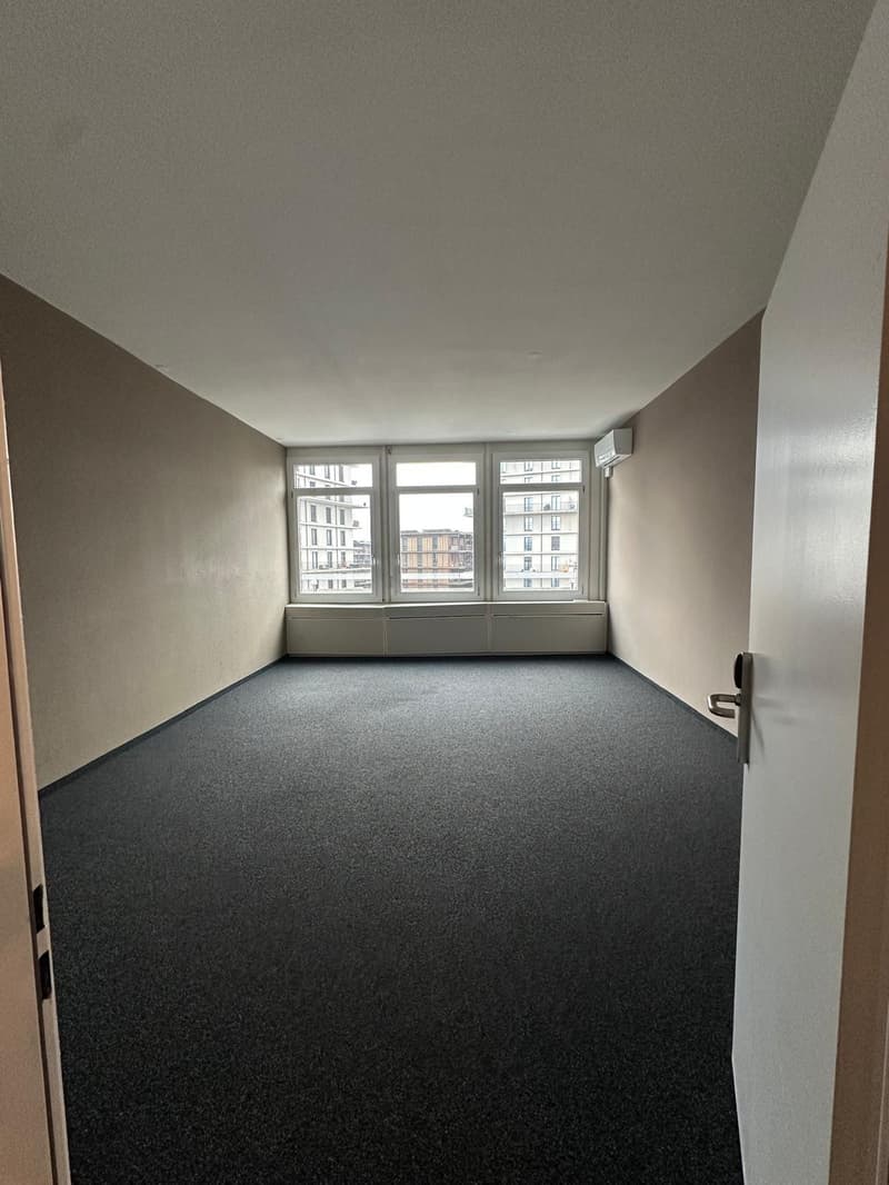 Large room with gray walls, gray carpeted floor, and large windows overlooking buildings outside.