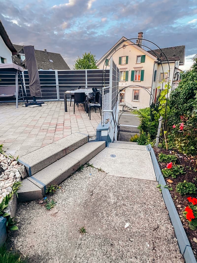 Outdoor dining area with table and chairs, brick patio, steps leading to the entrance, garden with flowers and plants, metal fence, house in the background