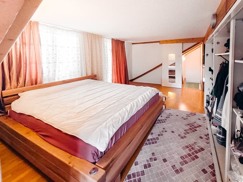 Bedroom with wooden bed, white and red bedspread, wooden floor, white walls, large window with curtains, wooden wardrobe, rug, wooden staircase