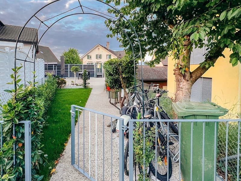 A garden with a gravel pathway, bicycles parked, a tree, and a trash bin.