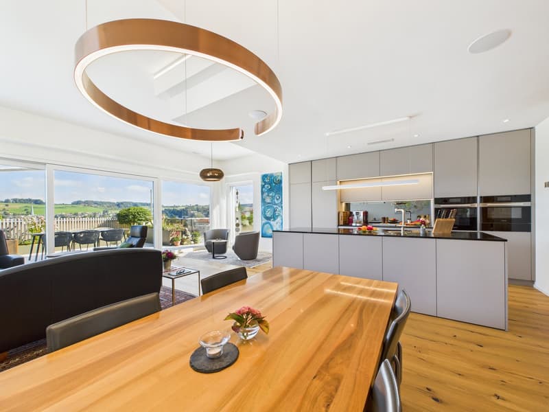 Living and dining area with wooden floor, gray cabinets, central wooden table with flowers, large windows, black sofas, and a circular lamp.