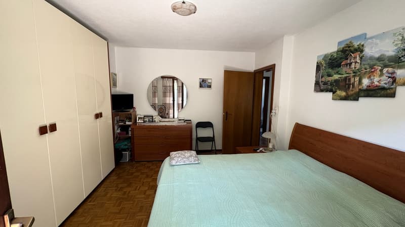 The image shows a bedroom with a double bed, a wooden dresser with a round mirror, and a TV stand. The room has white walls and a parquet floor. There are also some artwork and decorative items visible.