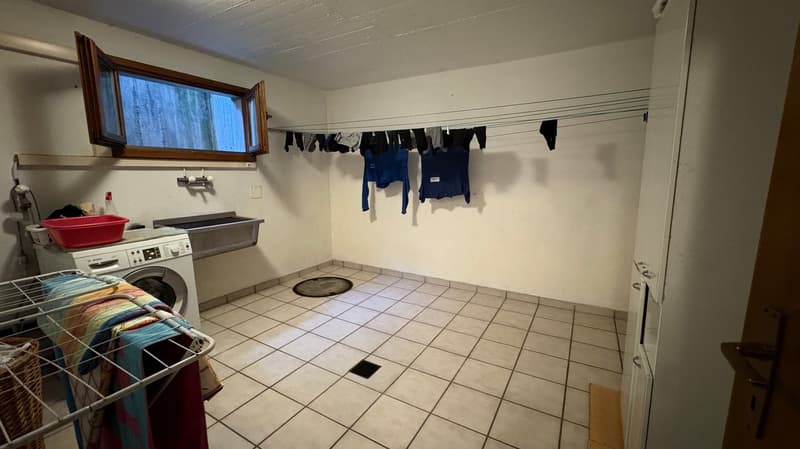 Laundry room with a washing machine, drying rack, and clothes hanging on a line. The room has tile flooring and a window.