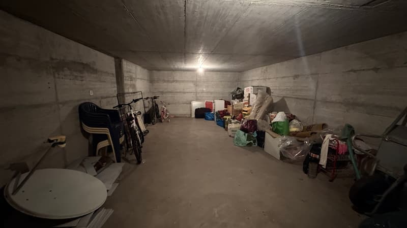 The image shows a cluttered storage room with various items such as chairs, boxes, and other miscellaneous objects stored in the space. The room has concrete walls and ceiling, giving it a utilitarian and industrial appearance.
