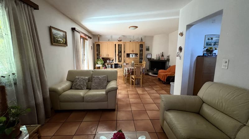 Spacious living room with leather sofas, wooden cabinets, framed artwork, and a fireplace. The room has tile flooring and large windows providing natural light.