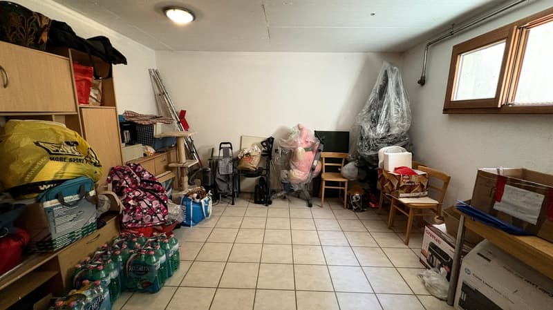 The image shows a cluttered storage room or basement with various items such as boxes, bags, furniture, and other household items stored in the space. The room has a tiled floor and appears to be used for storage purposes.