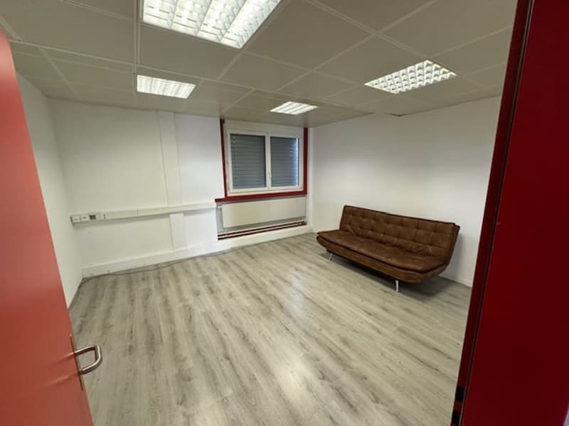 The image shows a spacious office space with a modern and minimalist design. The room has a hardwood-style flooring, white walls, and recessed lighting fixtures. There is a brown leather sofa placed in the corner, providing a comfortable seating area. The