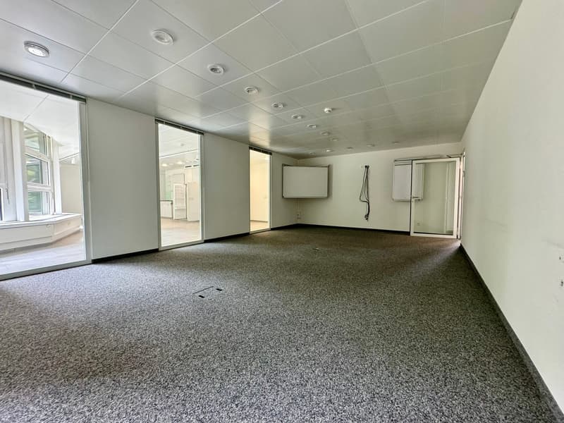 The image shows a large, open office space with a tiled ceiling, recessed lighting, and a carpeted floor. The room has several windows along one wall, providing natural light. There is a projection screen mounted on the wall, and some electrical outlets a