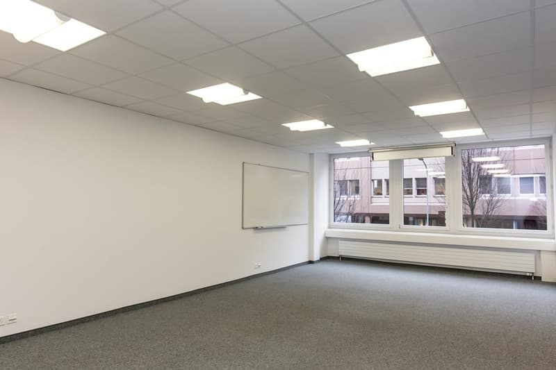 This image depicts an empty office space with white walls, a large window, and a whiteboard mounted on the wall. The room is equipped with recessed lighting and has a carpeted floor. The space appears to be suitable for use as a conference room, meeting r