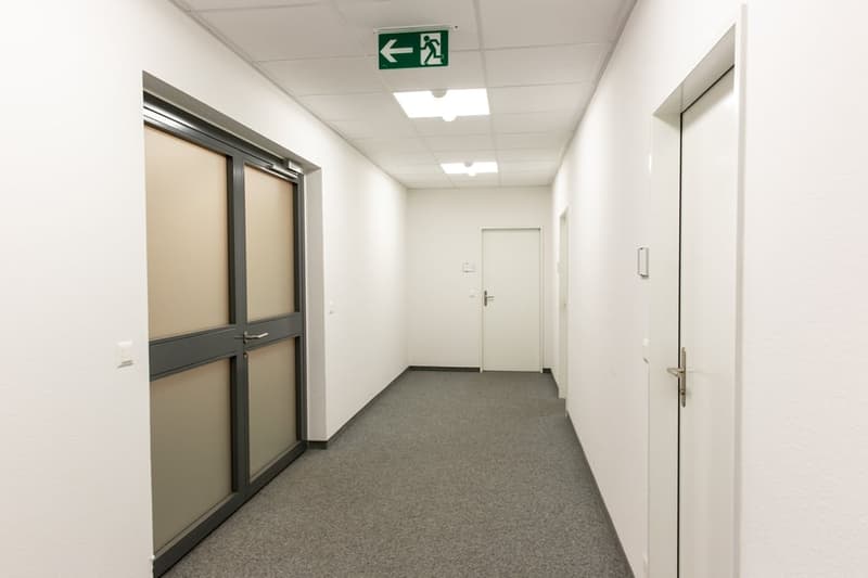 The image shows a long, well-lit office hallway with white walls, a gray carpeted floor, and a green exit sign on the wall. The hallway has several doors leading to different rooms or offices, and the lighting fixtures on the ceiling provide a bright and