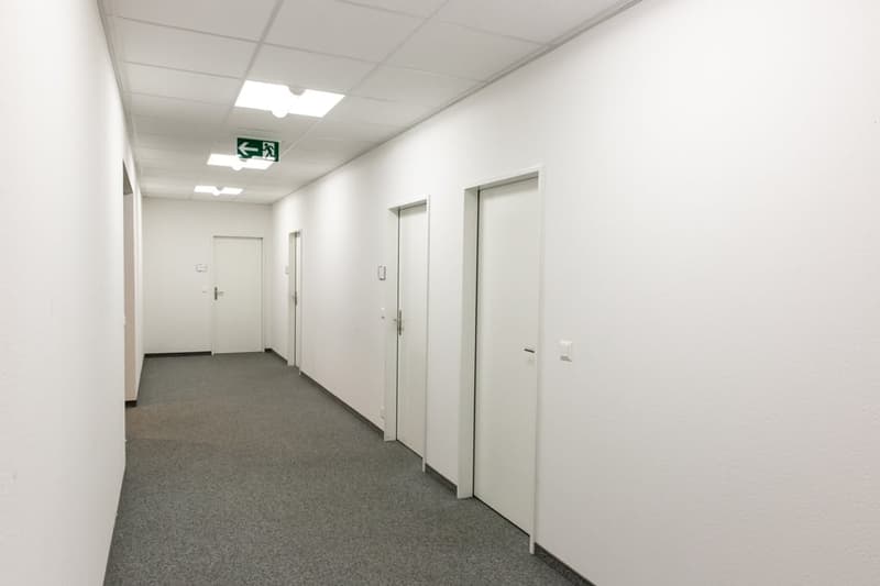 The image shows a long, narrow hallway with white walls and a gray carpeted floor. There are several white doors lining the hallway, and an exit sign can be seen on the wall. The lighting in the hallway appears to be provided by recessed ceiling lights.