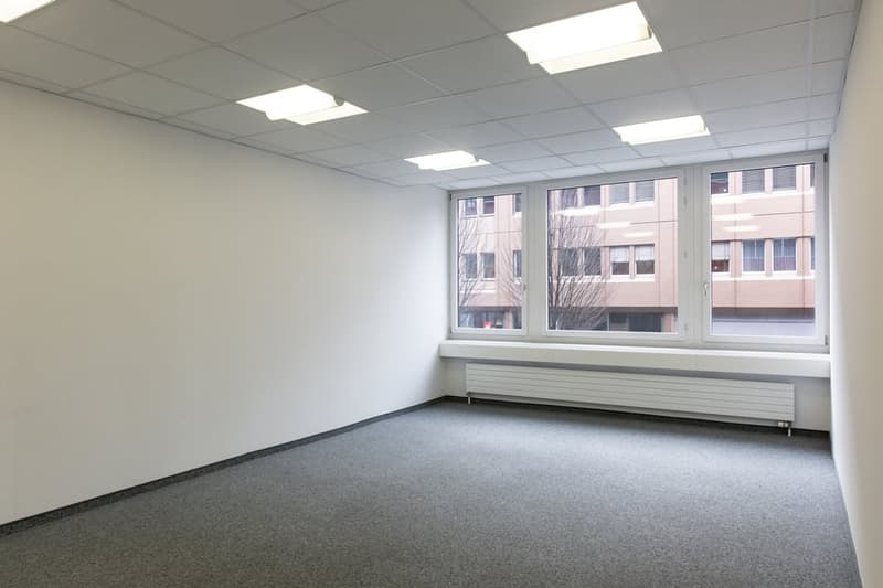 Empty office space with white walls, gray carpeted floor, and large windows overlooking other buildings.