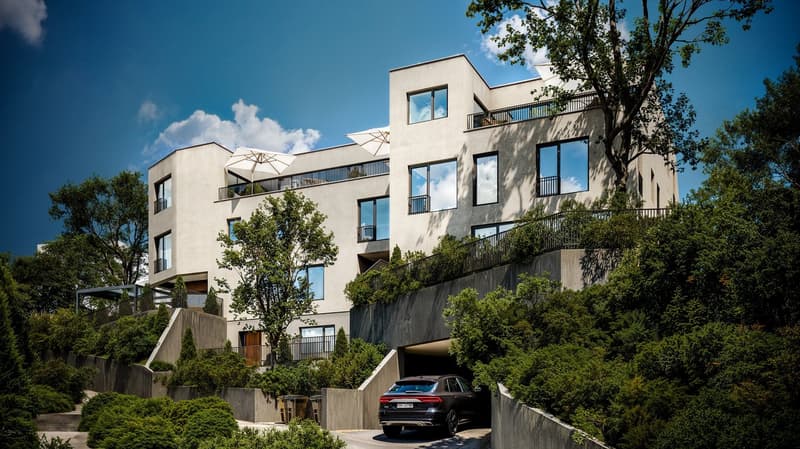 Multi-story apartment building with balconies, surrounded by trees and greenery, with a parking area in the foreground