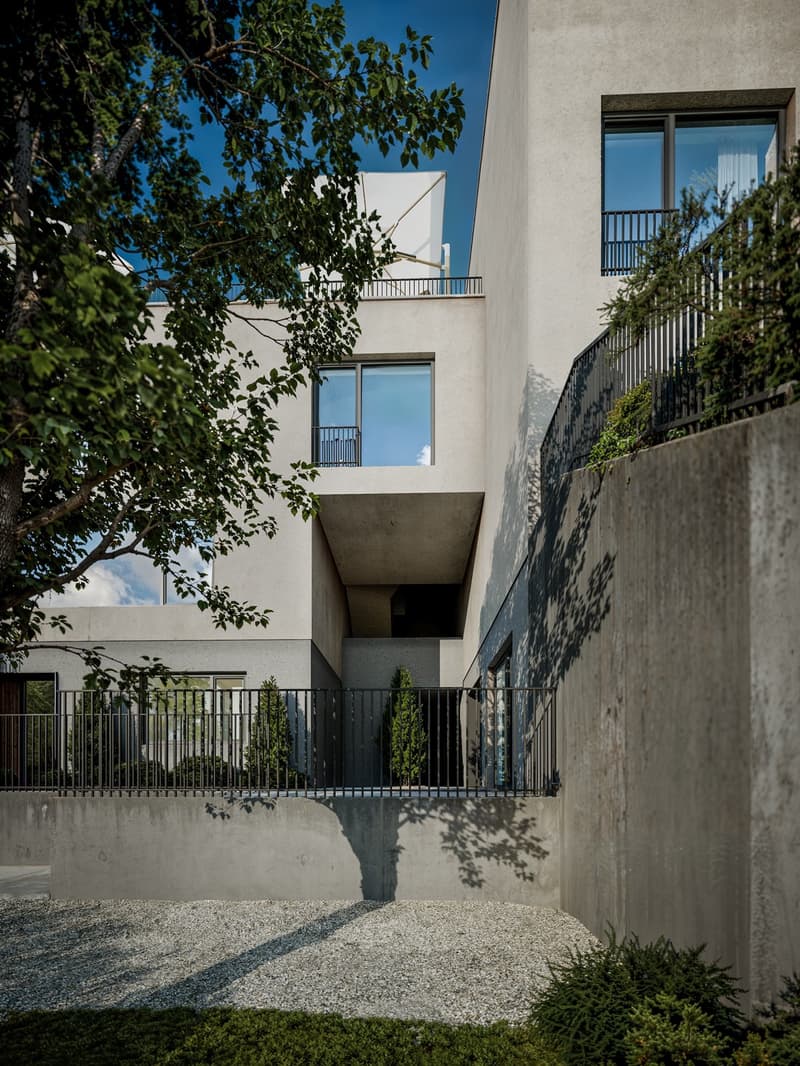 Multi-story residential building with white exterior walls, large windows, and balconies. The building is surrounded by trees and has a gravel pathway leading to the entrance.