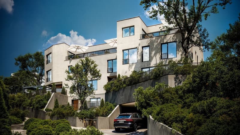 Multi-story apartment building with balconies, surrounded by trees and greenery, with a parking area in the foreground