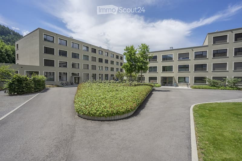 Multi-story apartment building with gray exterior, large windows, and a landscaped courtyard with greenery and a paved walkway. There are bicycle racks visible, indicating parking space for residents.