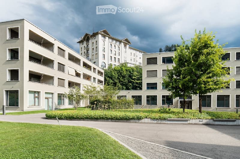 Multi-story apartment building with balconies, surrounded by green landscaping and trees, with a paved walkway in the foreground.