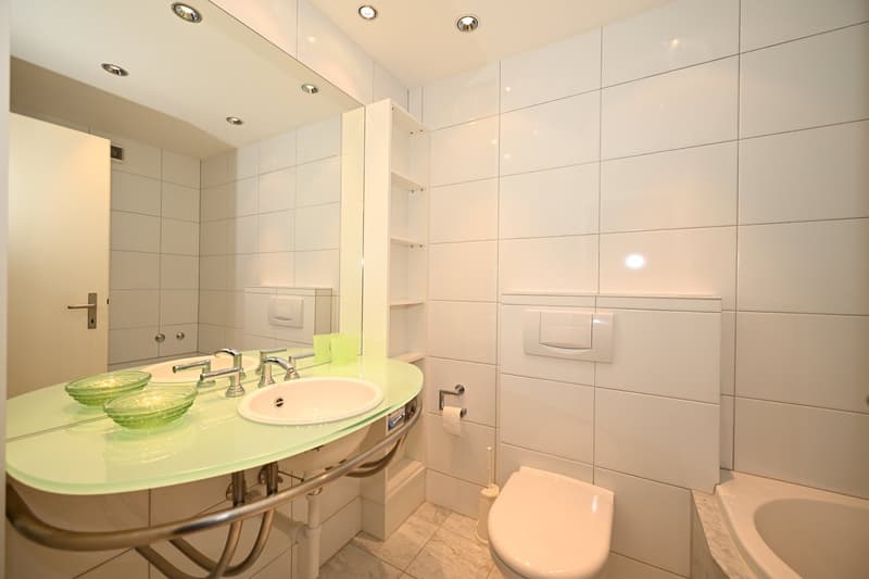 Bathroom with white tiles, green countertop, double sink, toilet, bathtub, mirror, shelves, and recessed lighting.