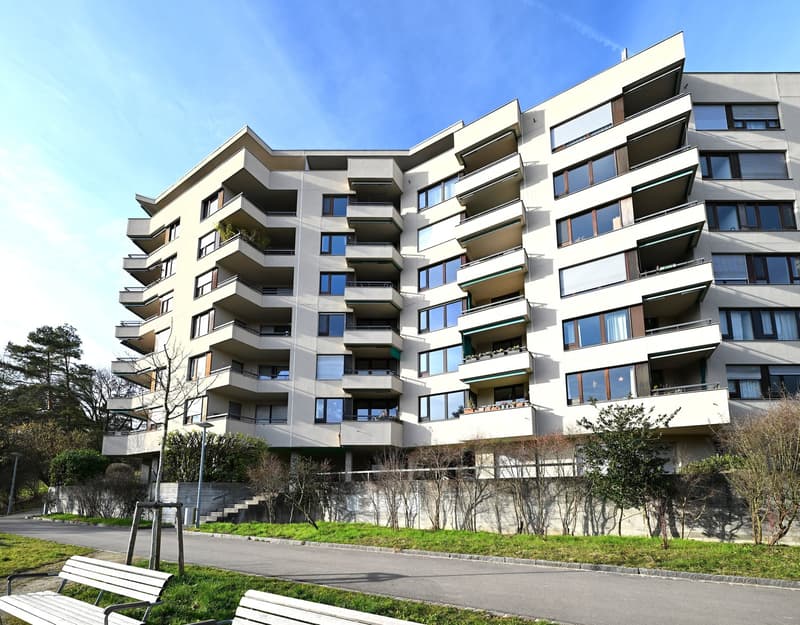 Modern multi-story apartment building with balconies, white exterior, large windows, and a landscaped area in front.