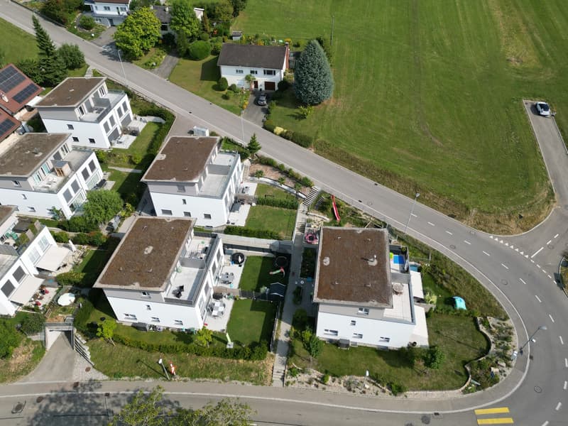 Aerial view of a residential area with several houses, driveways, and a road. The houses are white with brown roofs, and some have solar panels. The area is surrounded by green grass and trees, with a large open field in the background.