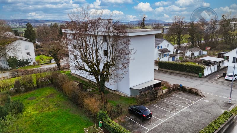 White building, 3 stories, parking lot, car parked, green grass, trees, bushes, road