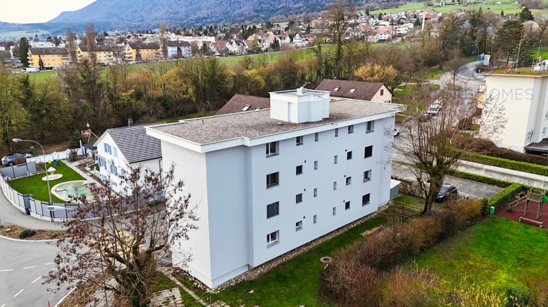 Aerial view of a white apartment building with multiple windows, a small garden, and a fountain in front. The building is surrounded by trees and grass, with a road and a car parked nearby. In the distance, there are more buildings and a mountain range.