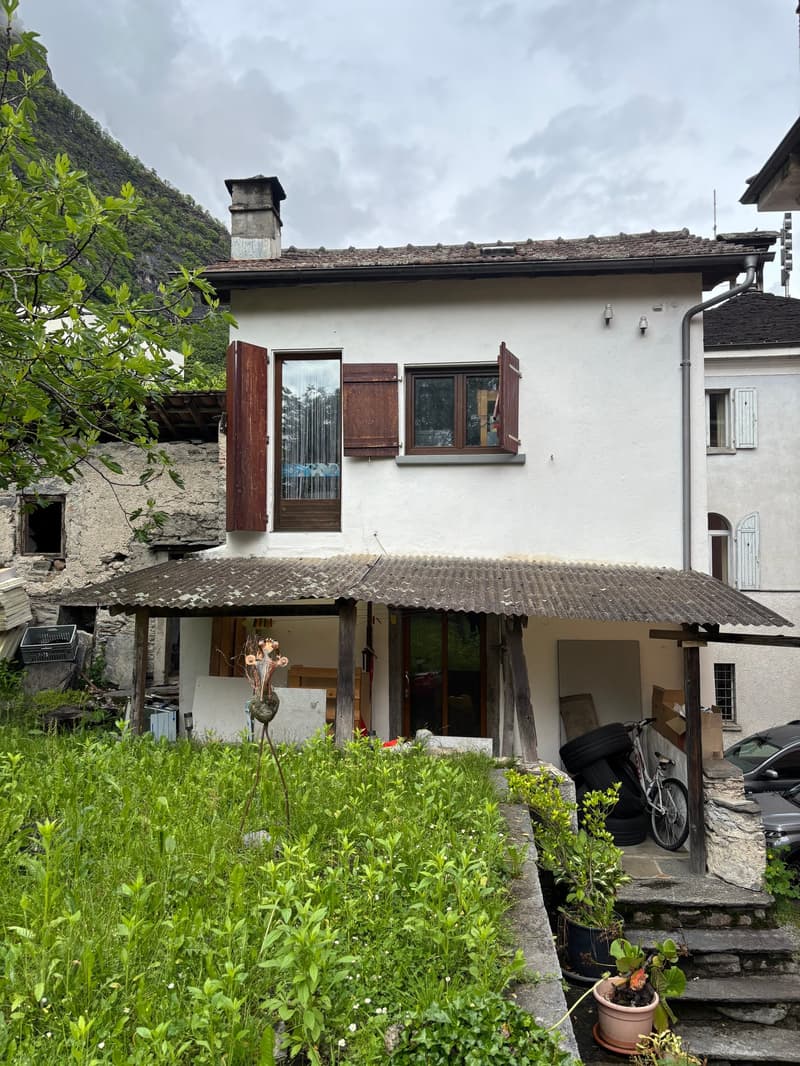 Two-story house with white walls, brown wooden shutters, and a tiled roof. There is a small balcony or terrace on the upper level. The house is surrounded by lush greenery and vegetation, with a bicycle parked in the foreground.