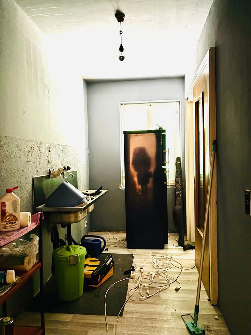 Cluttered room with various household items and appliances, including a green container, a black cabinet, and electrical cords on the floor.