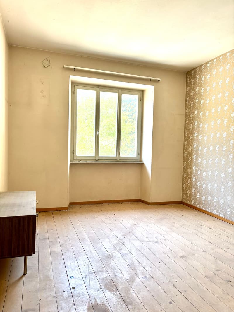 Empty room with beige walls, a large window, and a wooden floor. The room appears to be in need of renovation, with some damage visible on the floor.