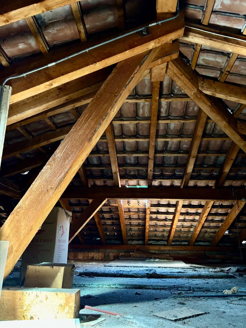 The image shows the interior of an attic space with a wooden truss roof structure and shelves or storage units lining the walls. The floor appears to be made of wooden boards and there are various items and debris scattered throughout the space.