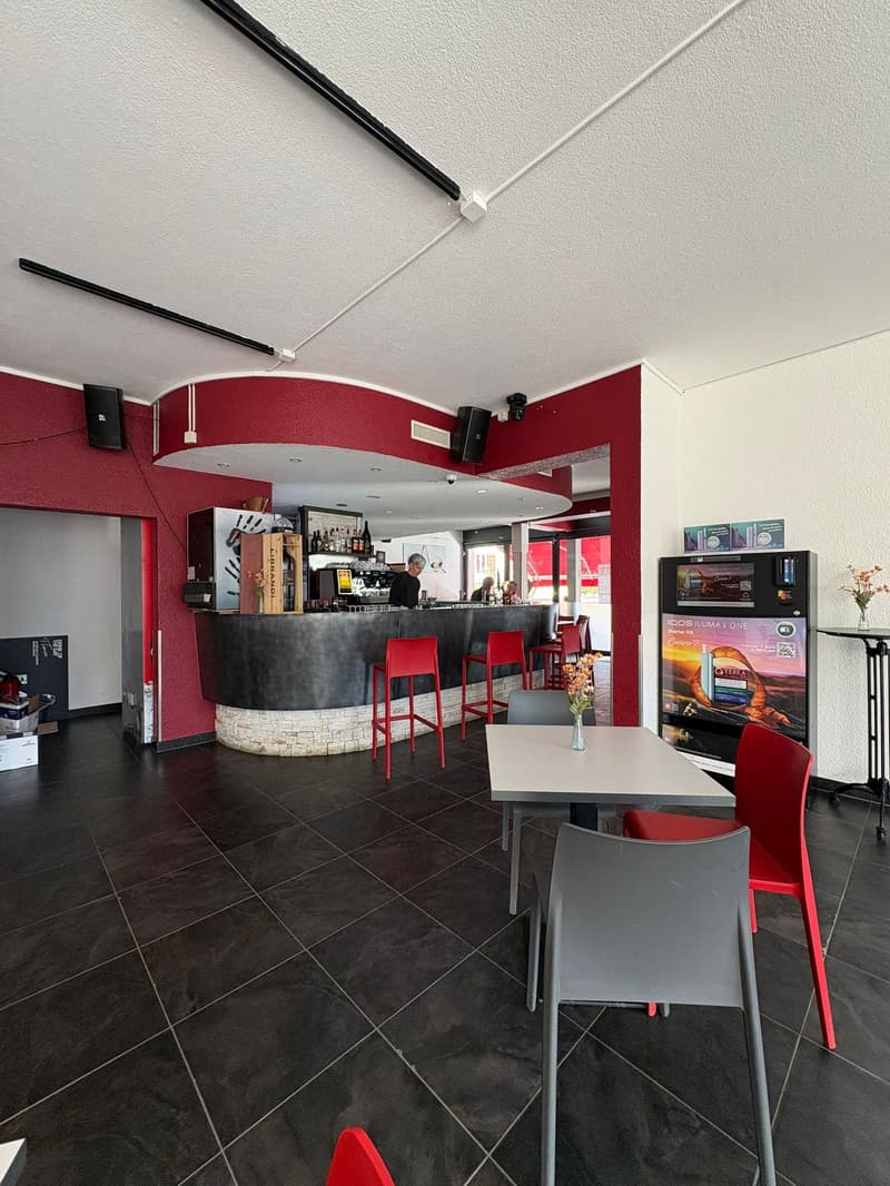 A bar area with a black tiled floor, a red-painted L-shaped counter, gray and red chairs, a small round table, and an advertisement machine.