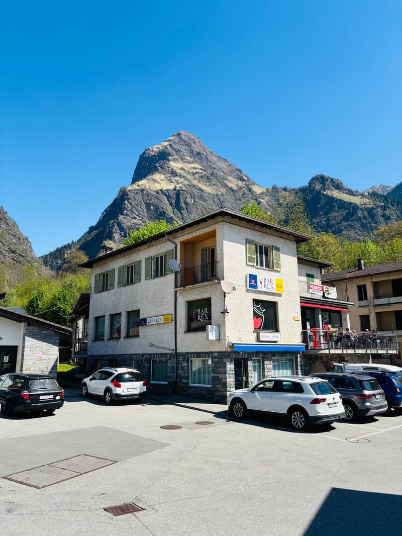 A multi-story building with a bar and restaurant on the ground floor, surrounded by parked cars and with a large mountain in the background.