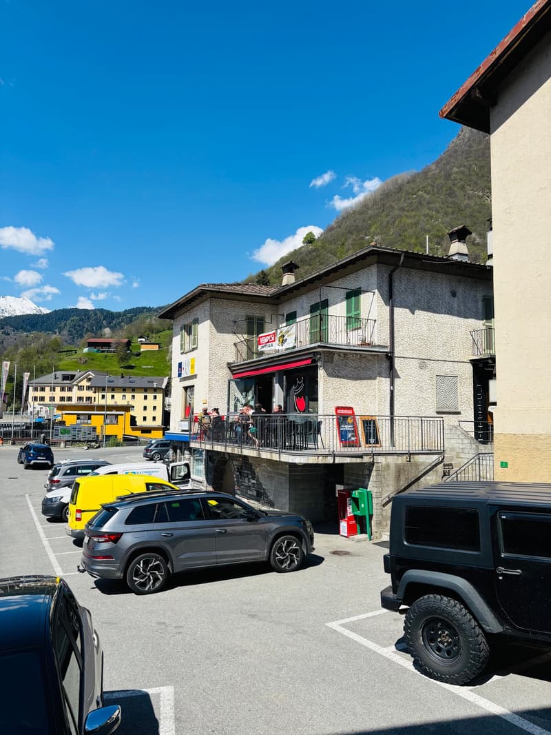 The image shows a street view of a small town or village in a mountainous area. There are several buildings with balconies and shops on the ground floor. There are various vehicles parked on the street, including a yellow taxi, a grey SUV, and a black off