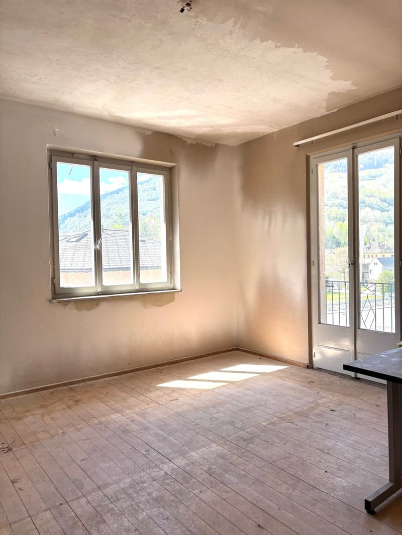 The image shows an empty room with a damaged ceiling, wooden floor, and large windows overlooking a scenic outdoor view. The room appears to be in need of renovation, with bare walls and a generally unfinished appearance.