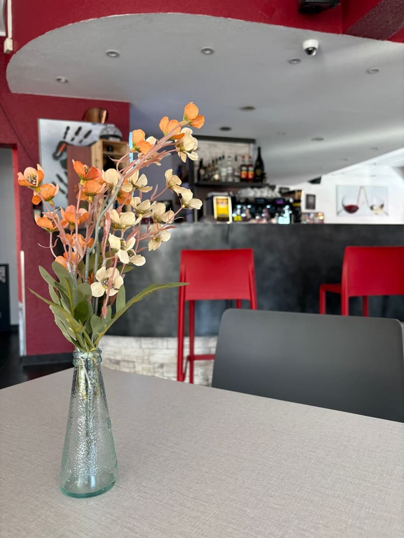 A clear glass vase with orange and white flowers on a dining table with a gray chair in the front, a red chair at the counter, a bar with bottles behind the counter, a white ceiling, a red wall, and a surveillance camera on the ceiling.