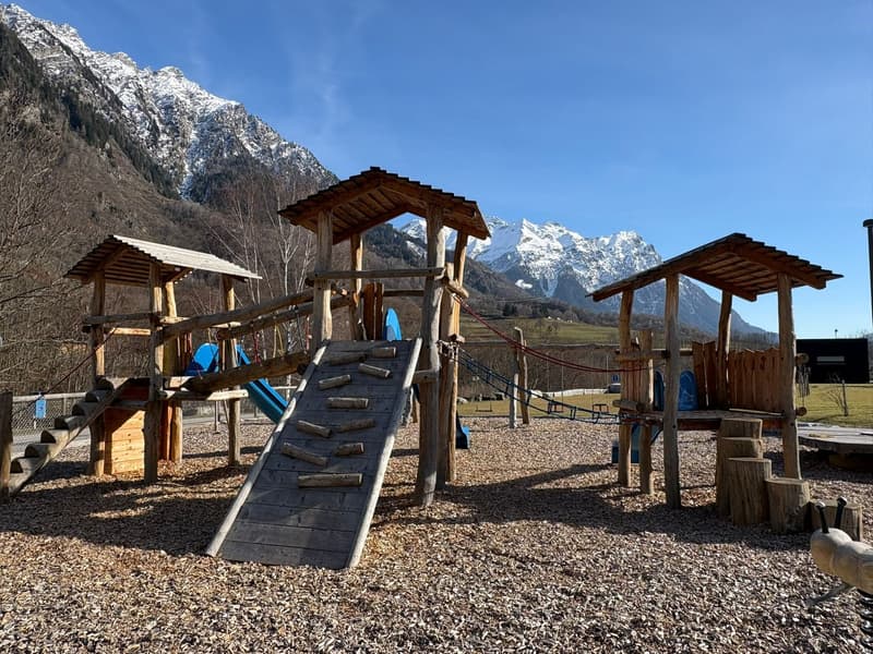wooden playground, climbing frame, slides, mountain backdrop, gravel ground